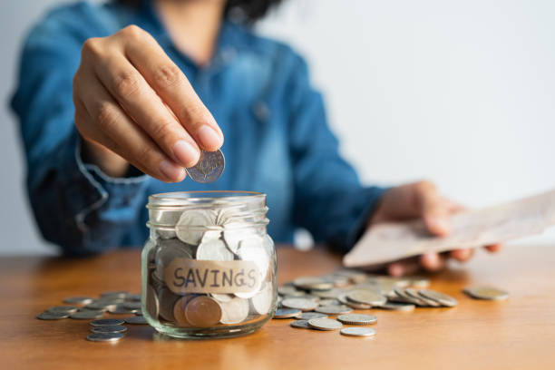 The Woman Hand Is Putting a Coin In a Glass Bottle And a Pile of Coins on a Brown Wooden Table